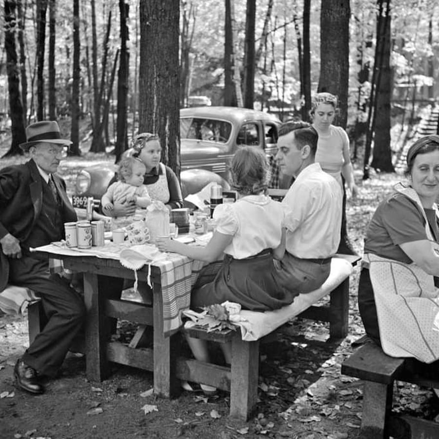A black-and-white photo of a family having a picnic at a wooden table in a wooded area, with an old car parked nearby. Adults and children sit and stand around the table, sharing food and drinks.