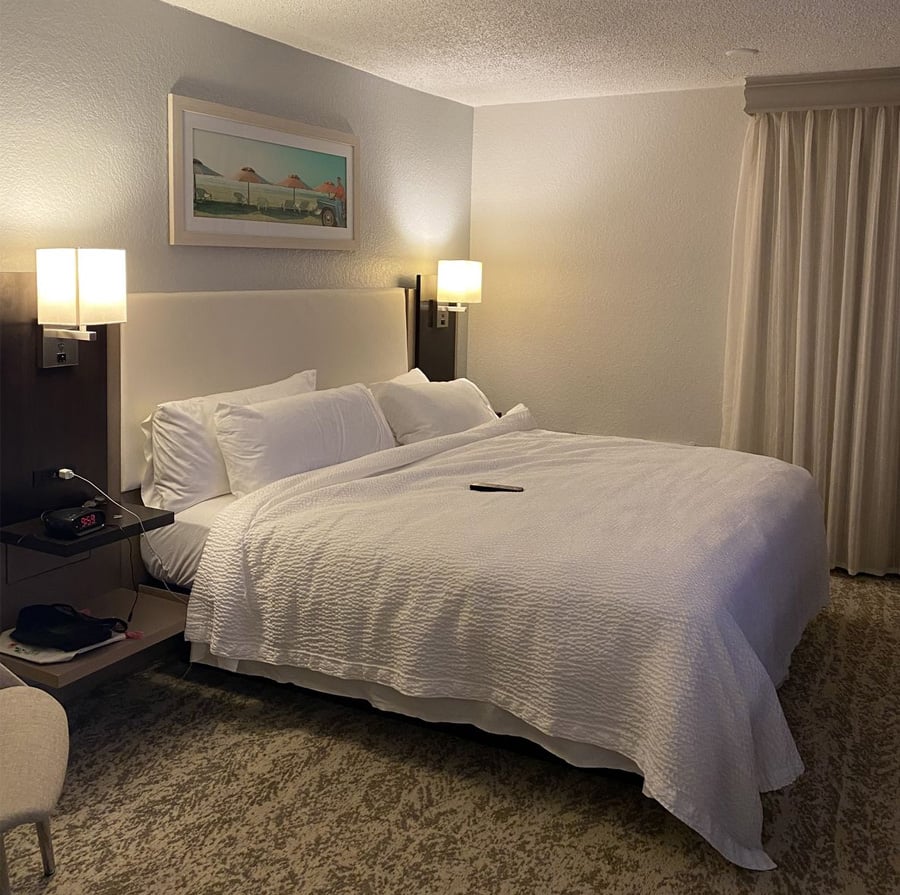A neatly made hotel bed with white linens and pillows, bedside shelves each with a lamp, a remote control on the bed, a framed picture above the headboard, and closed curtains on the right side.