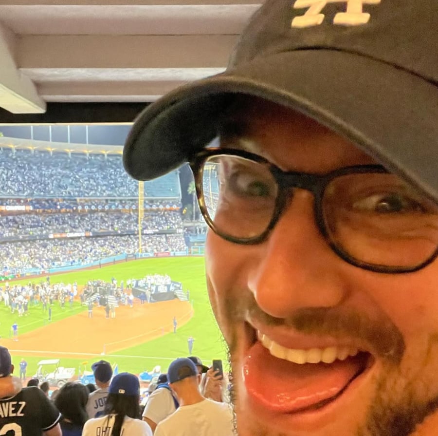 A smiling person in glasses and a baseball cap poses in the foreground with a crowded baseball stadium and a celebration on the field in the background.