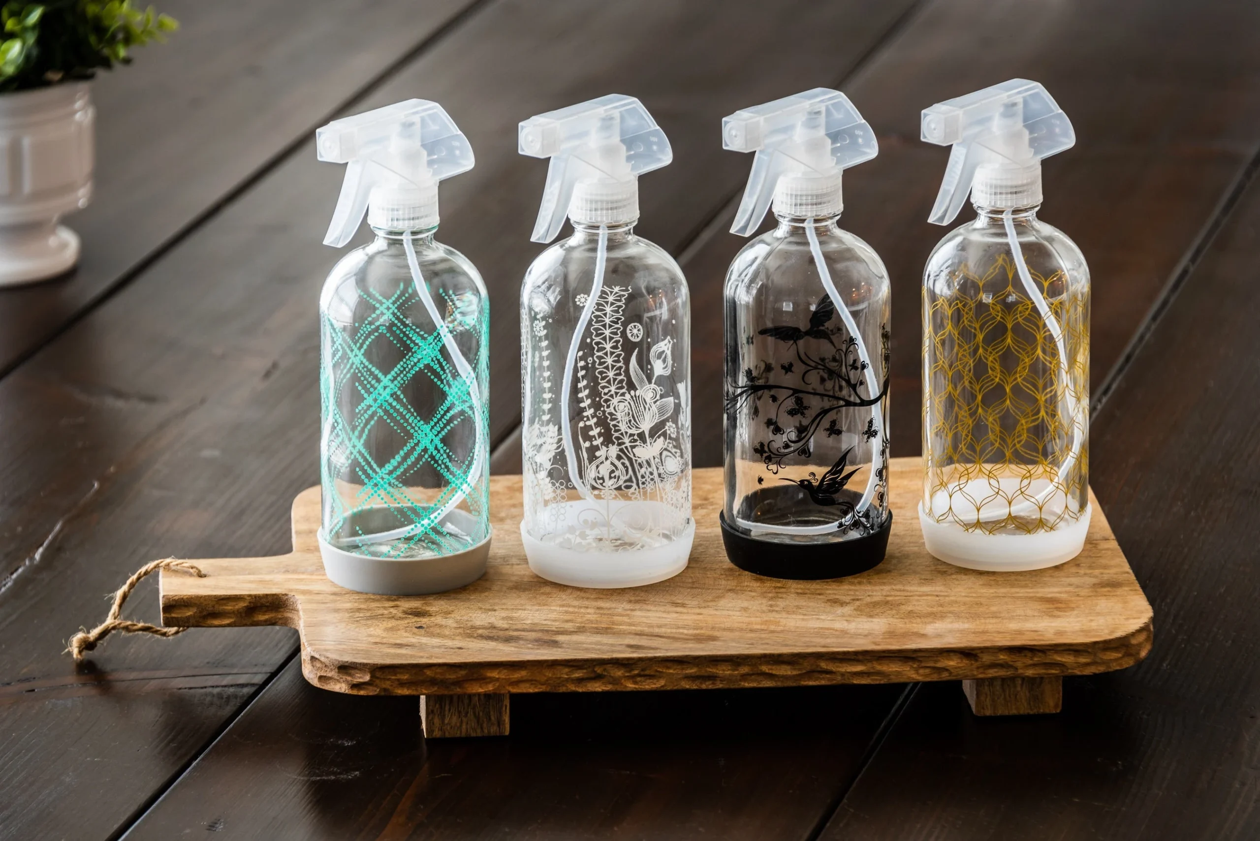 Four clear spray bottles with decorative patterns and different colored bases are arranged in a row on a wooden cutting board, positioned on a dark wooden table. A small plant is blurred in the background.