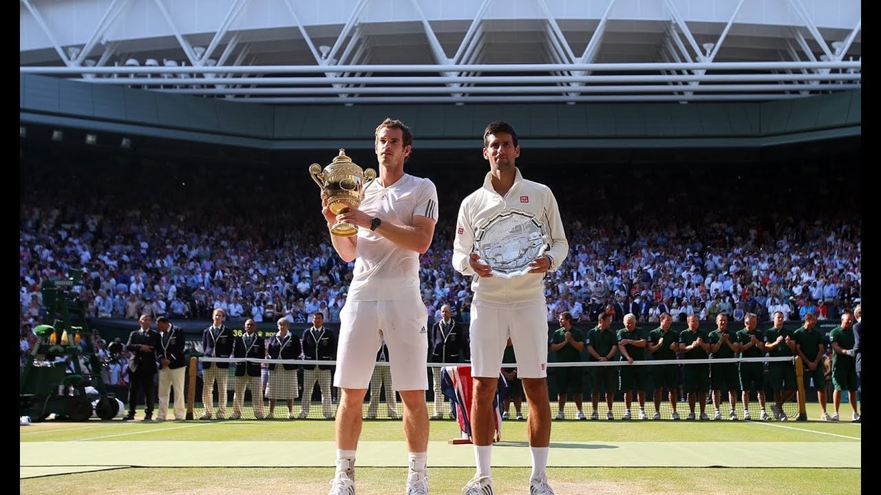 Two male tennis players stand on a grass court holding trophies, with one holding a gold cup and the other a silver plate. A large crowd fills the stadium behind them. Both are dressed in white sportswear.