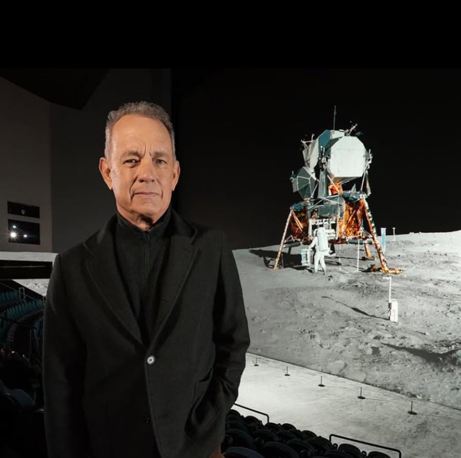 A man in a dark suit stands inside a theater or museum, with a large backdrop showing an astronaut on the moon near a lunar module. The setting appears to be an exhibition or presentation about space exploration.