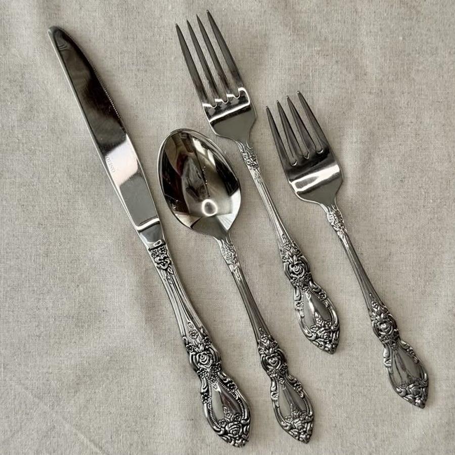 Four pieces of ornate silverware—a knife, spoon, dinner fork, and salad fork—are arranged on a light fabric background. Each utensil has an intricate floral design on the handle.