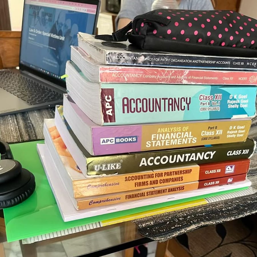 A stack of accountancy and financial textbooks is piled on a glass table next to a laptop, headphones, and some folders, indicating a study or work environment.