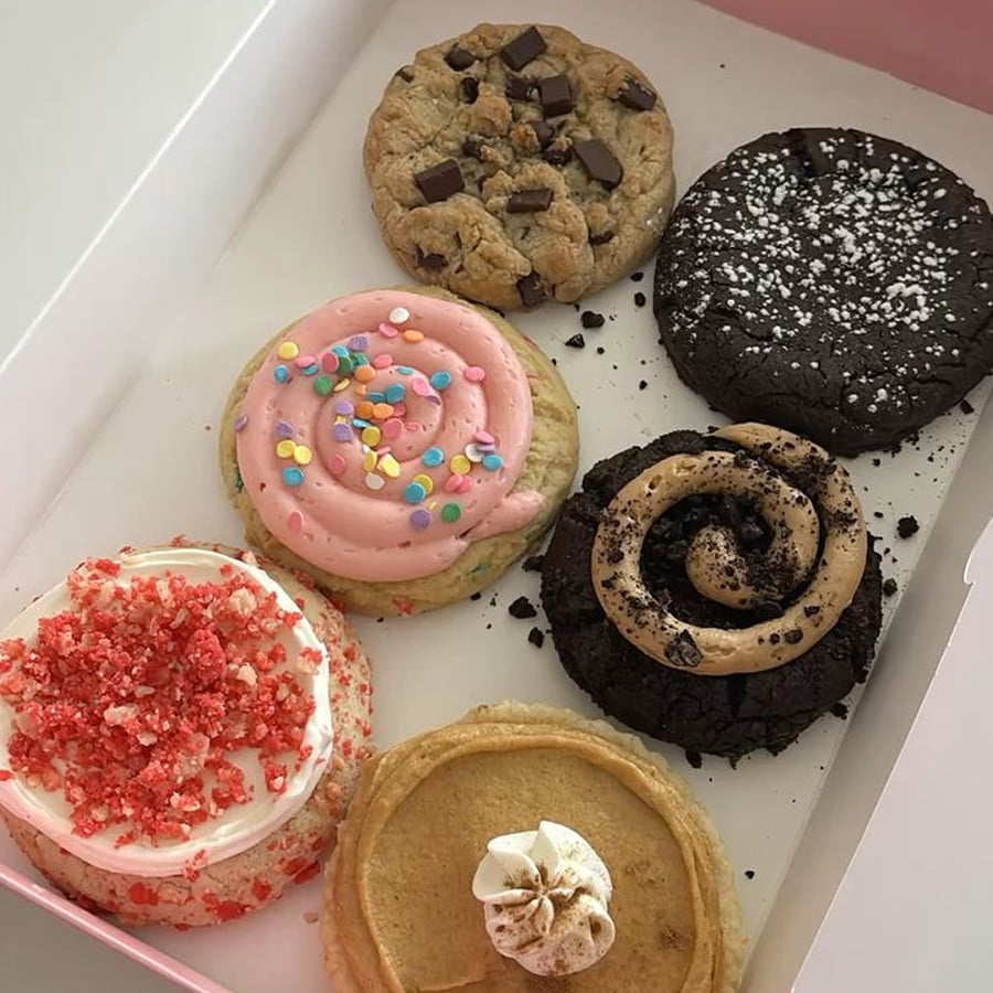 A box with six large cookies, each with different toppings: chocolate chunks, powdered sugar, pink icing with sprinkles, white icing with red crumbs, swirled frosting, and a swirl of white cream.