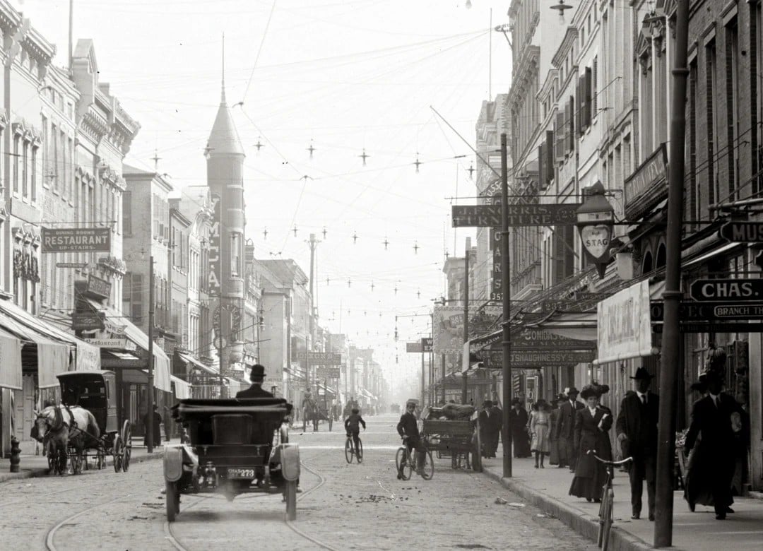 A busy city street scene from the early 1900s with people walking, riding bicycles, horse-drawn carriages, vintage cars, and storefronts lining both sides under a web of overhead wires.
