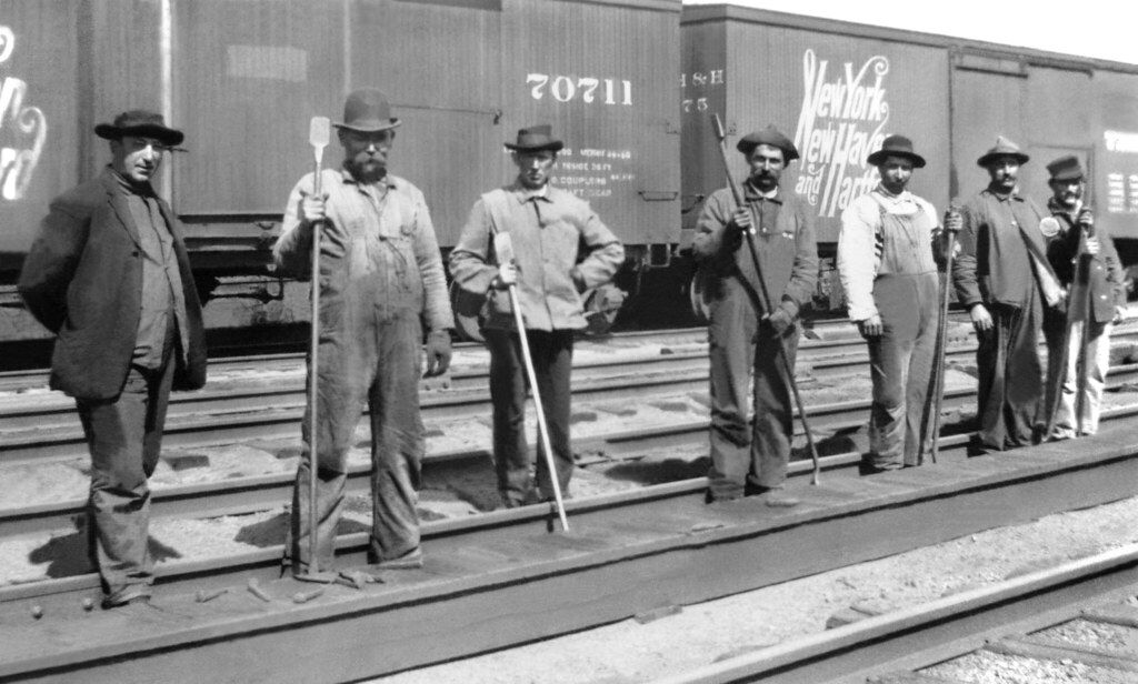 Seven railroad workers in work clothes and hats stand side by side on train tracks, holding tools, with train boxcars visible in the background. The image appears to be historical and in black and white.