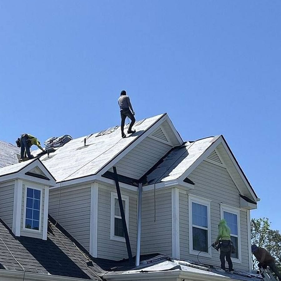 Several workers are installing or repairing roofing material on top of a two-story house under a clear blue sky. Some tools and materials are visible on the roof.