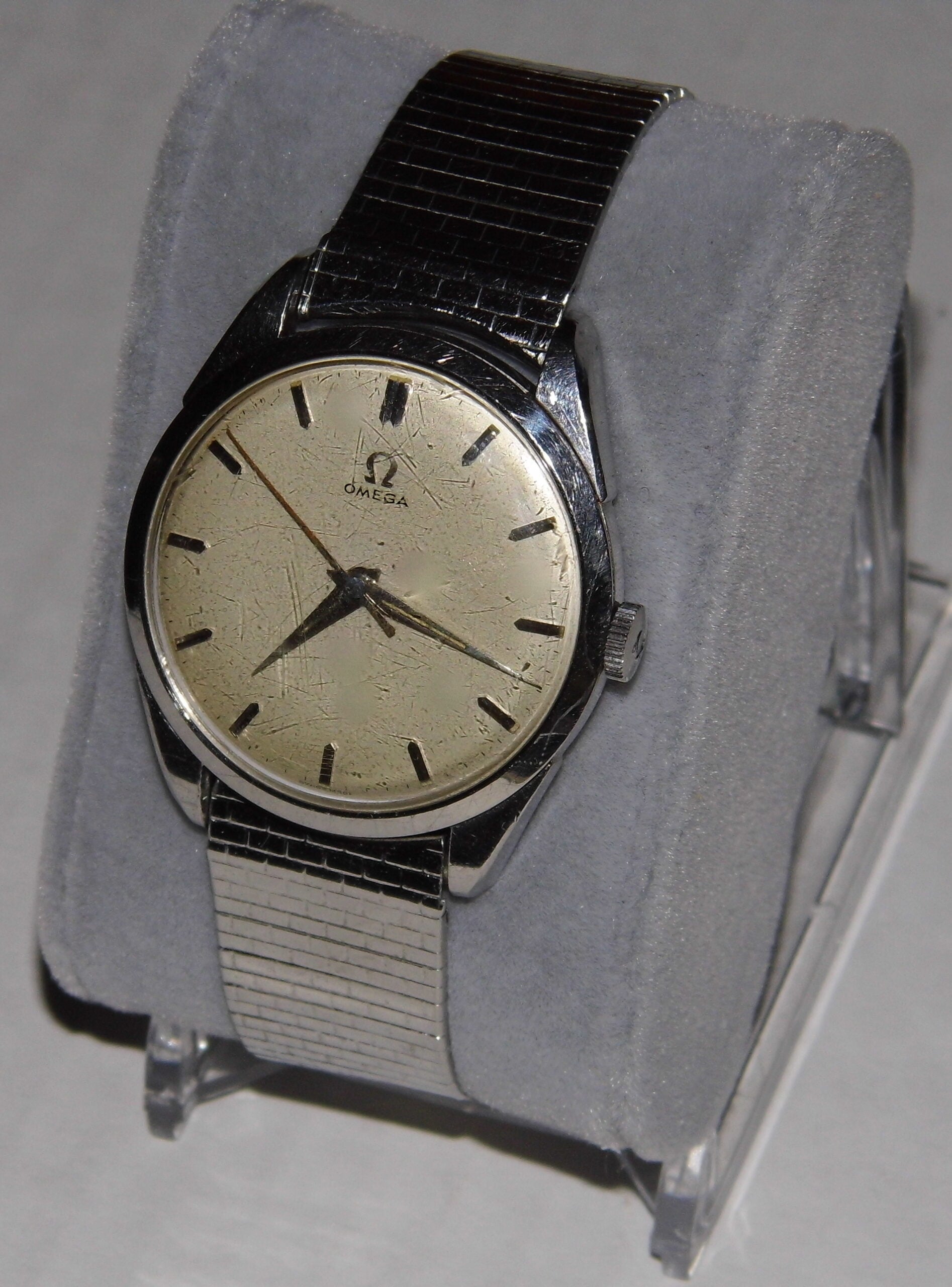A vintage Omega wristwatch with a metal band and a cream-colored, worn dial is displayed on a gray cushion stand. The watch has black hour markers and hands.