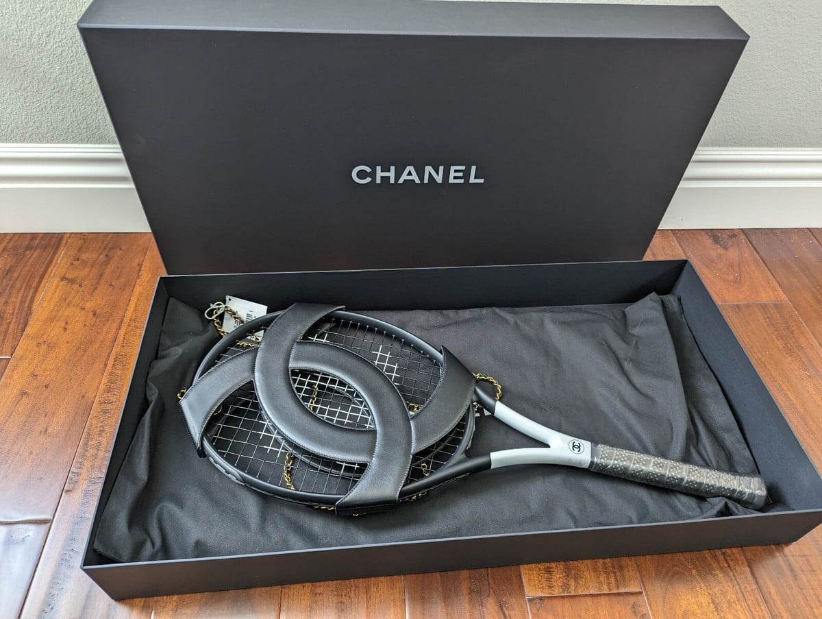 A black Chanel tennis racket with the iconic interlocking C logo, elegantly displayed in a black Chanel-branded box lined with black fabric on a wooden floor.