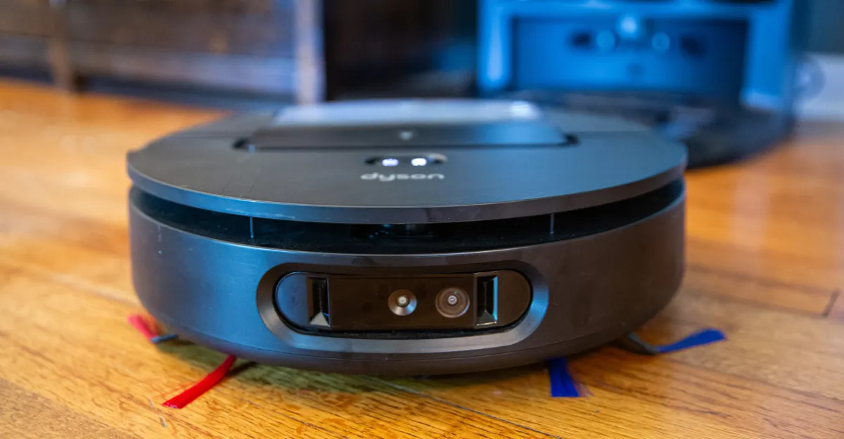 A black Dyson robot vacuum cleaner is shown on a wooden floor, with its camera and sensors visible on the front. Another similar device is blurred in the background.