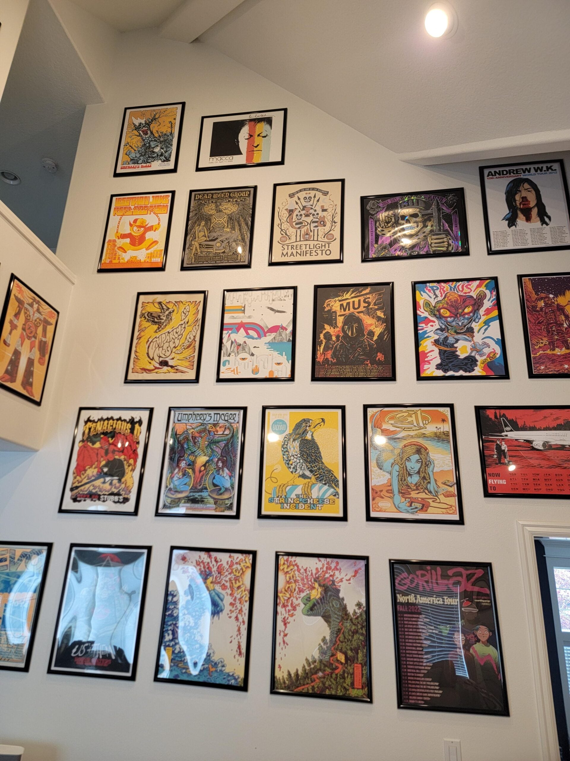 A white wall displays a grid of colorful framed posters, mostly concert or event art, arranged from floor to ceiling in a bright room with high ceilings and modern decor.