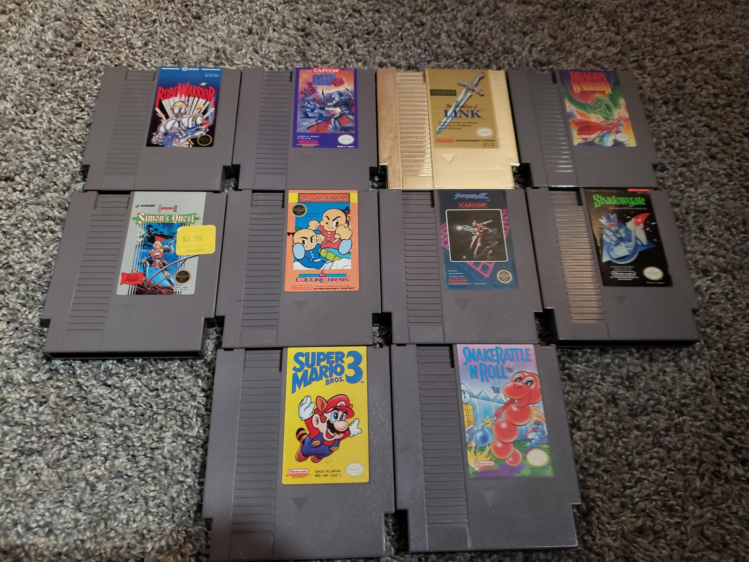 Nine NES game cartridges arranged in three rows on a gray carpet. Visible titles include Super Mario Bros. 3, The Legend of Zelda, Double Dragon, Snake Rattle N Roll, StarTropics, and a few others.