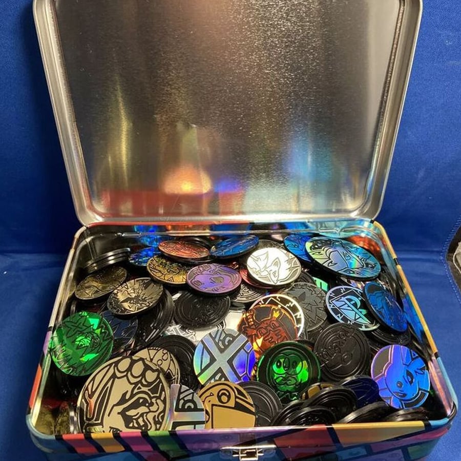 An open metal tin filled with colorful, shiny plastic tokens or coins featuring various cartoon characters and designs against a blue background.