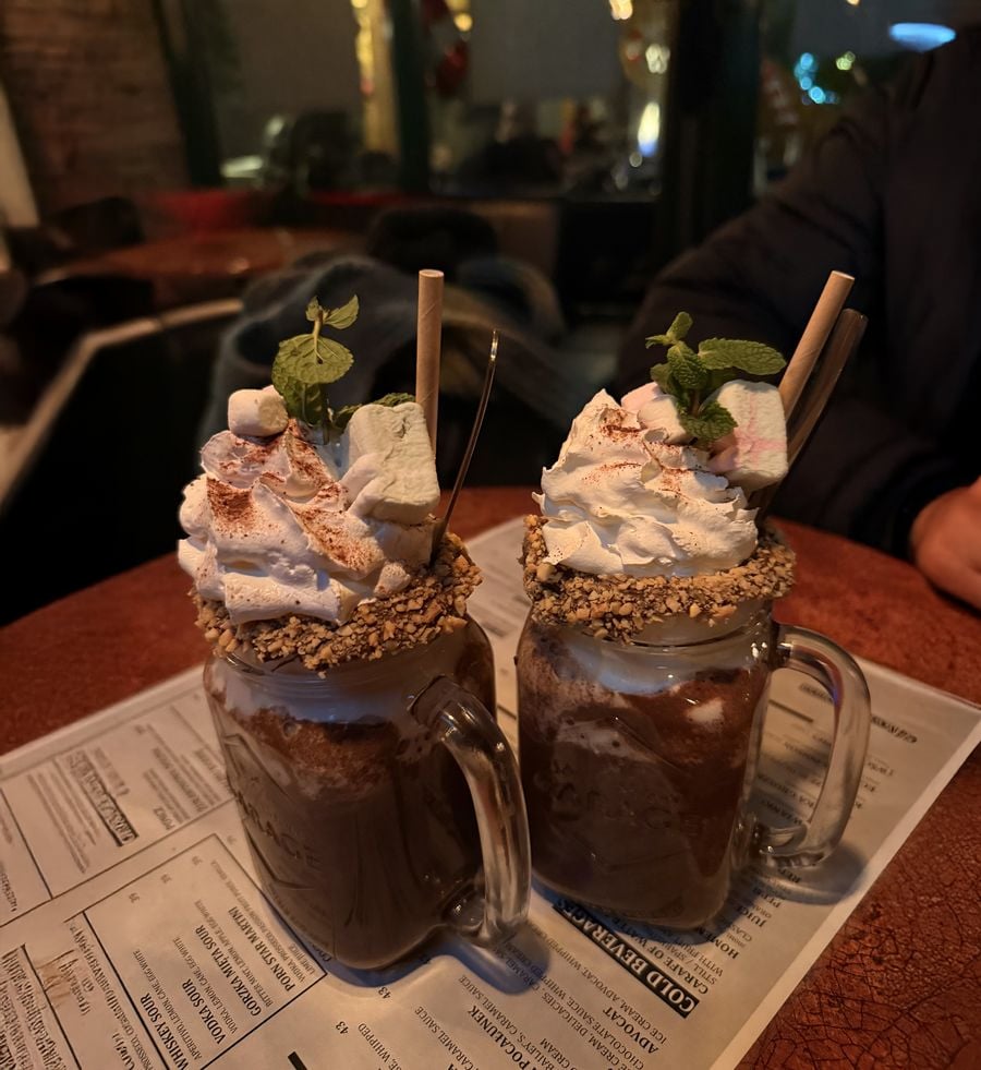 Two glass mugs filled with chocolate drinks topped with whipped cream, marshmallows, cocoa powder, mint leaves, and wafer straws, placed on a menu on a table in a cozy café setting.