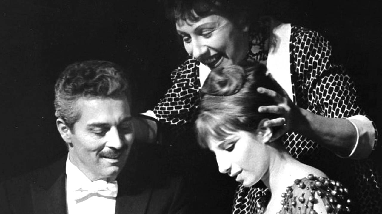 A man in a tuxedo sits next to a woman in a beaded dress looking down, while another woman stands behind them, smiling and placing her hands gently on the seated woman's head.