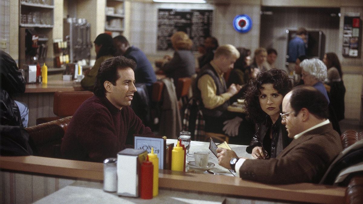 Three people sit at a booth in a busy diner, talking. The table has ketchup, mustard, and menus. Other diners are seated in the background. The atmosphere is warm and casual.
