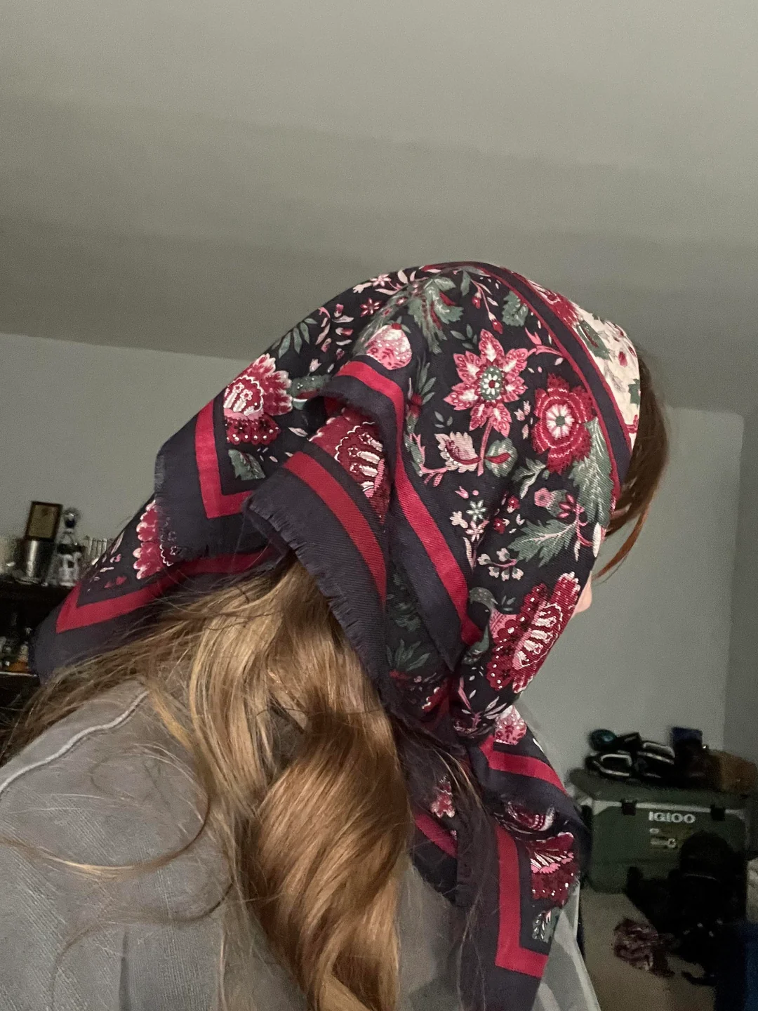 A person with long, wavy light brown hair wears a dark floral scarf with red, pink, and green patterns tied over their head. The background shows a room with shelves, boxes, and a green storage bin.