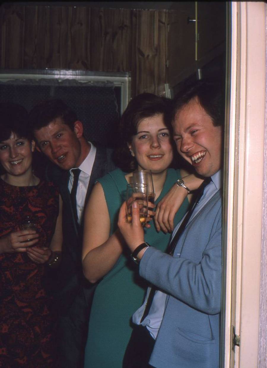 Four young adults stand closely together indoors at a party, smiling and holding drinks. Two women and two men are dressed in smart attire, appearing relaxed and happy in a warmly lit room.