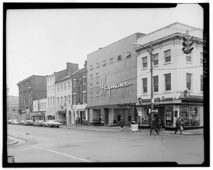 Black and white photo of a city street corner with storefronts, including "Jayman's" and "Pharmacy Shoe Store." Several people walk on the sidewalk and vintage cars are parked along the street.