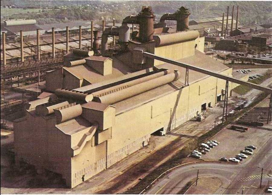 A large industrial factory building with multiple pipes and smokestacks, surrounded by parked cars and additional industrial structures in the background.