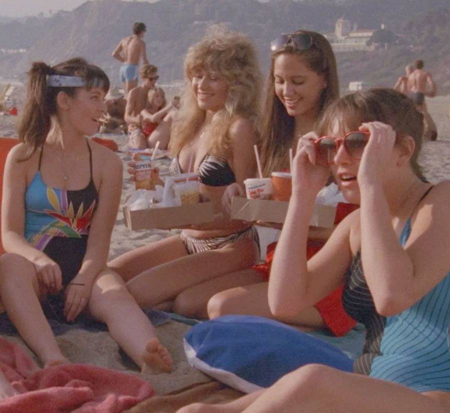 Four young women in swimsuits sit on the sand at a beach, talking and smiling. They have food and drinks, with other beachgoers and hills visible in the background under sunny skies.