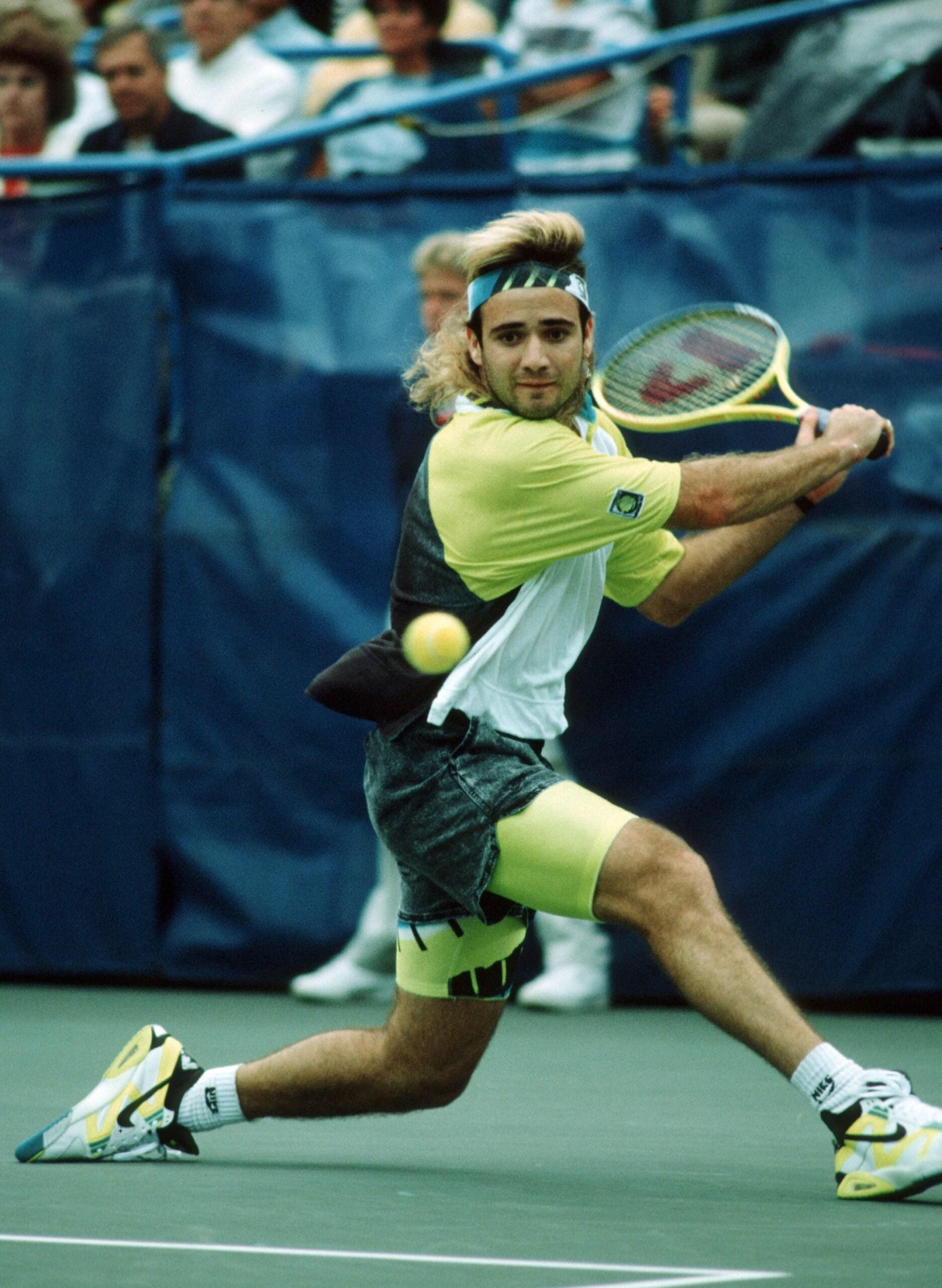 A tennis player with long hair and a headband prepares to hit a backhand shot during a match on an outdoor hard court, with spectators visible in the background.