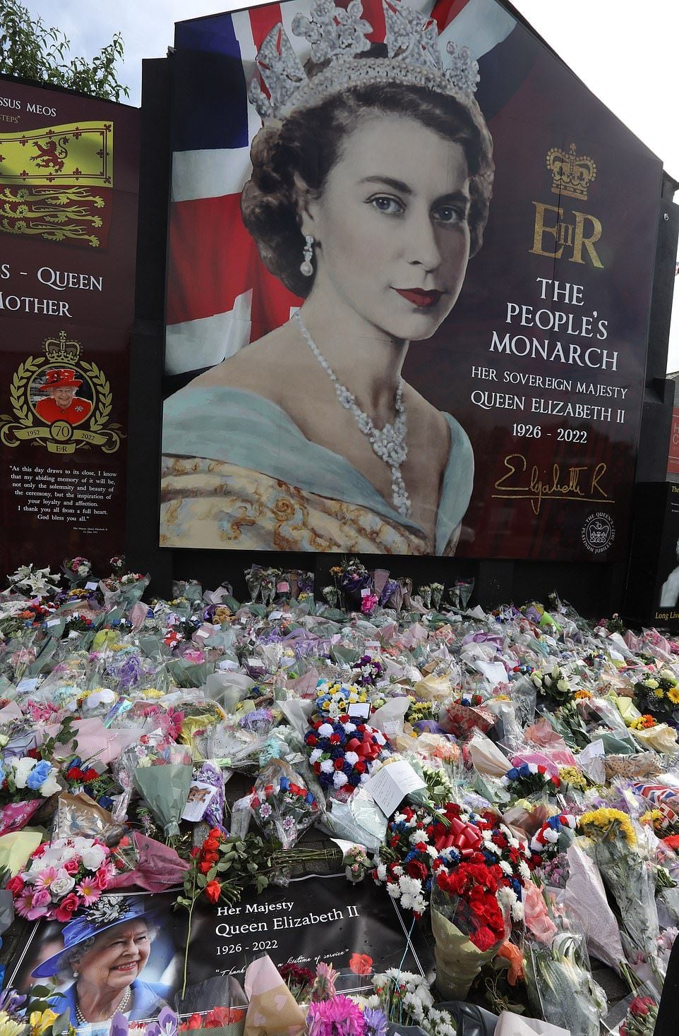 A large portrait of Queen Elizabeth II is displayed above numerous bouquets, cards, and tributes left by mourners, honoring her memory with messages and flowers after her passing in 2022.
