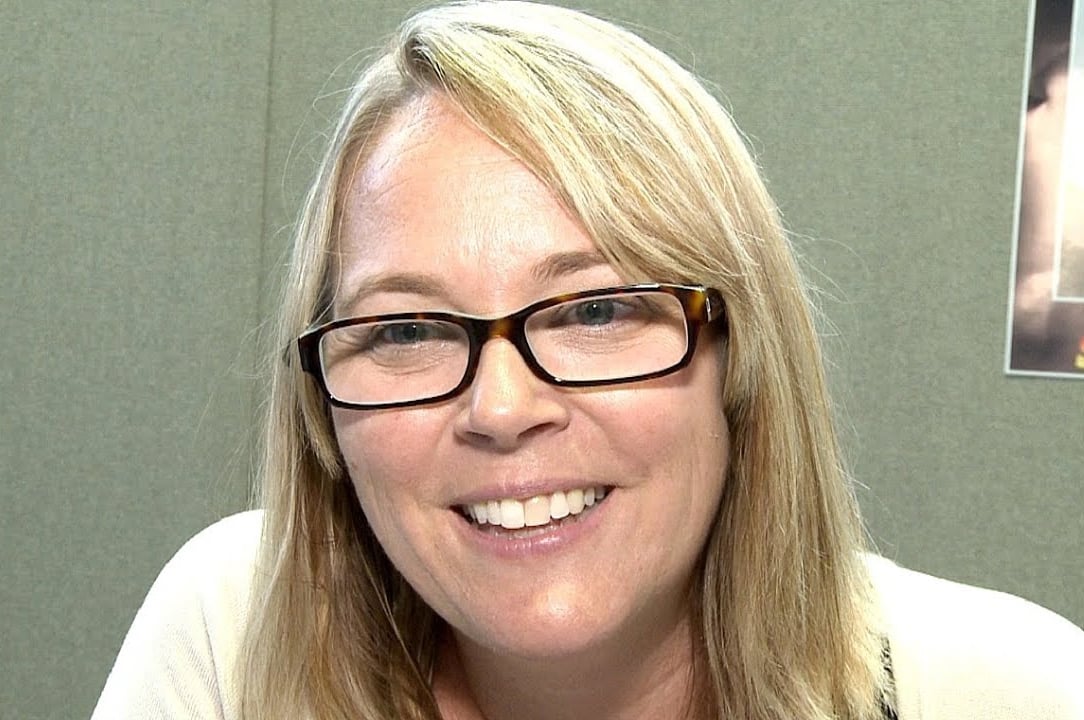 A woman with straight blonde hair and glasses smiles at the camera. She is indoors, in front of a plain gray wall, and is wearing a light-colored top.