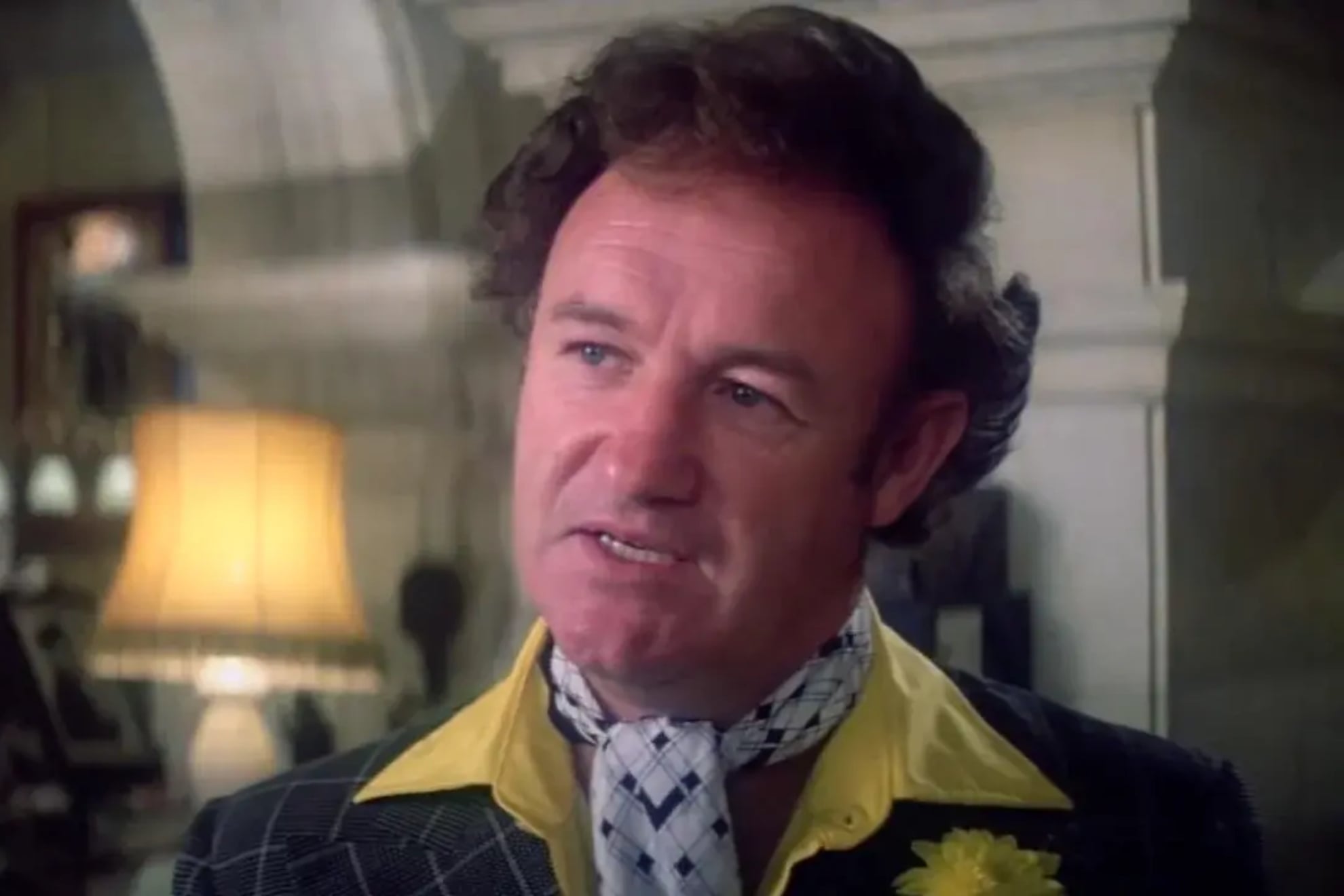 A man with curly brown hair wears a yellow-collared shirt, plaid jacket, and a patterned scarf around his neck, with a yellow flower pinned to his lapel. He stands indoors, lit by a lamp in the background.