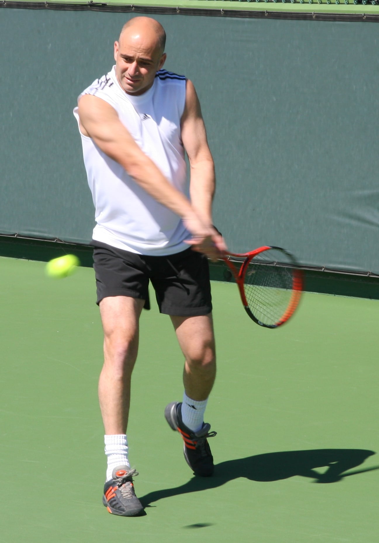 A man wearing a white sleeveless shirt and black shorts hits a tennis ball with a two-handed backhand on an outdoor tennis court.