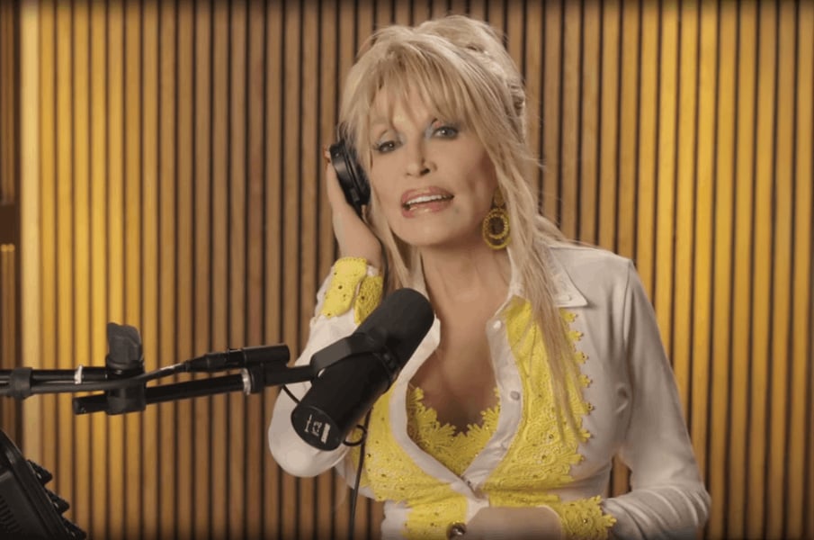 A woman with long blonde hair, wearing a white and yellow outfit, holds a headphone to her ear and speaks into a studio microphone in front of a wooden panel background.