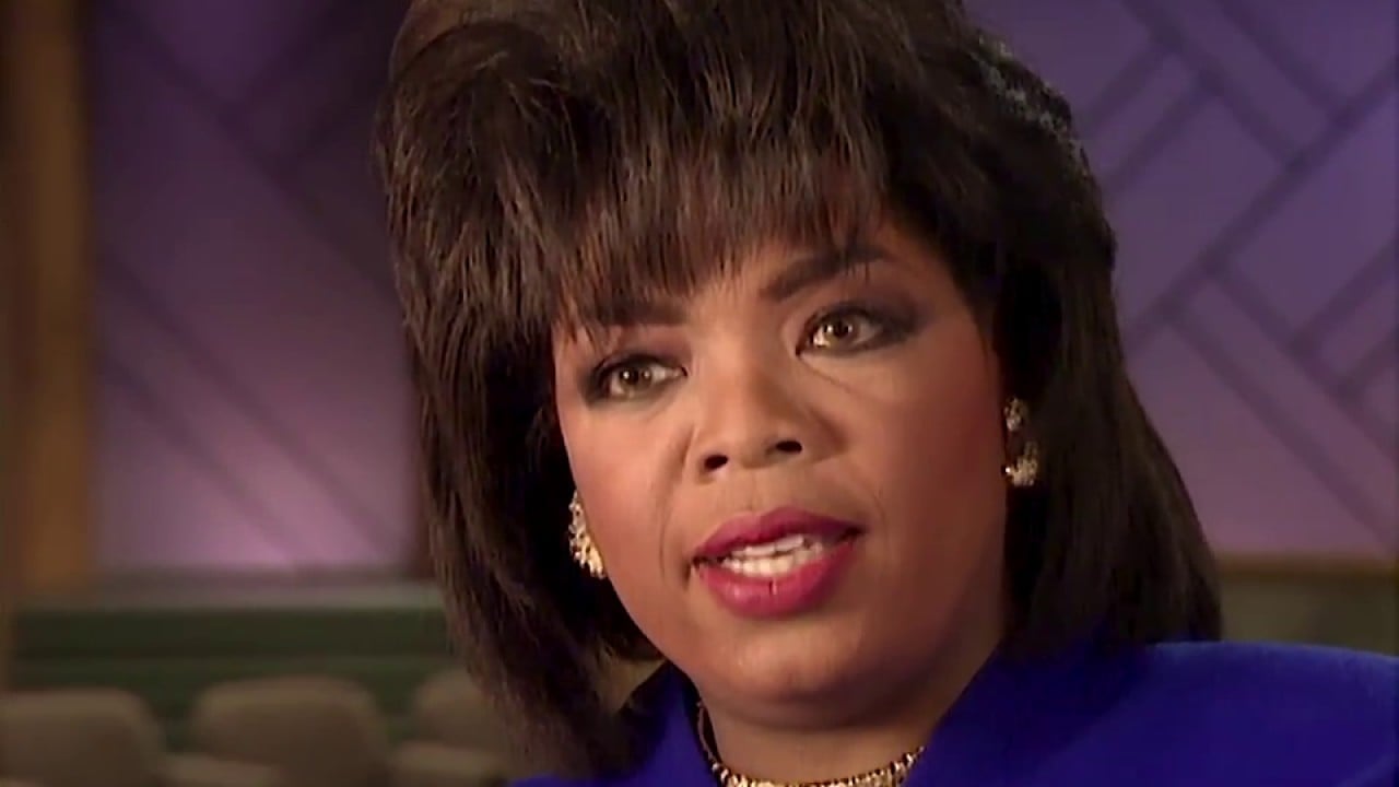A woman with short, styled dark hair is wearing gold earrings, a gold necklace, and a royal blue blazer. She is looking slightly to the side with a serious expression in a softly lit indoor setting.