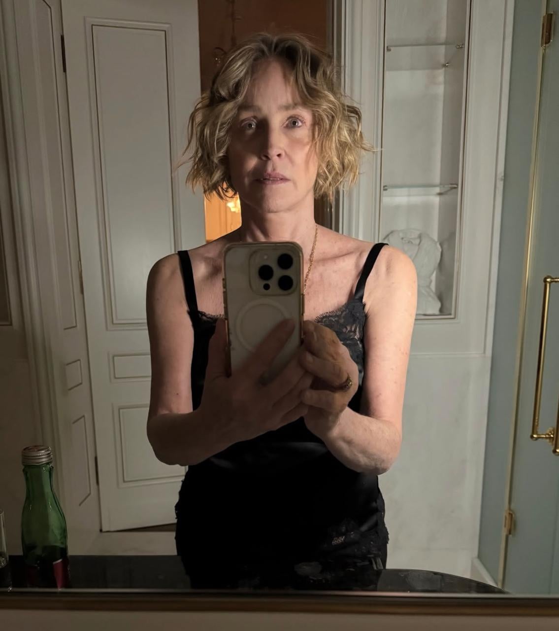 A woman with short, wavy blonde hair wears a black sleeveless top and takes a mirror selfie in a dimly lit bathroom with white paneling. A green glass bottle sits on the counter in front of her.