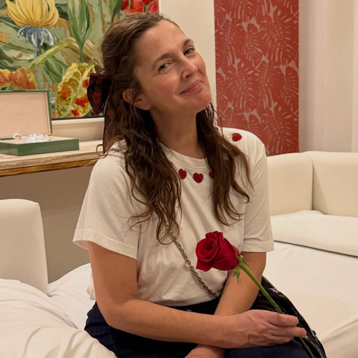 A woman with wavy brown hair sits on a white couch, smiling gently while holding a red rose. She wears a white t-shirt with red heart designs and a black ribbon in her hair. Colorful art and a jewelry box are visible in the background.