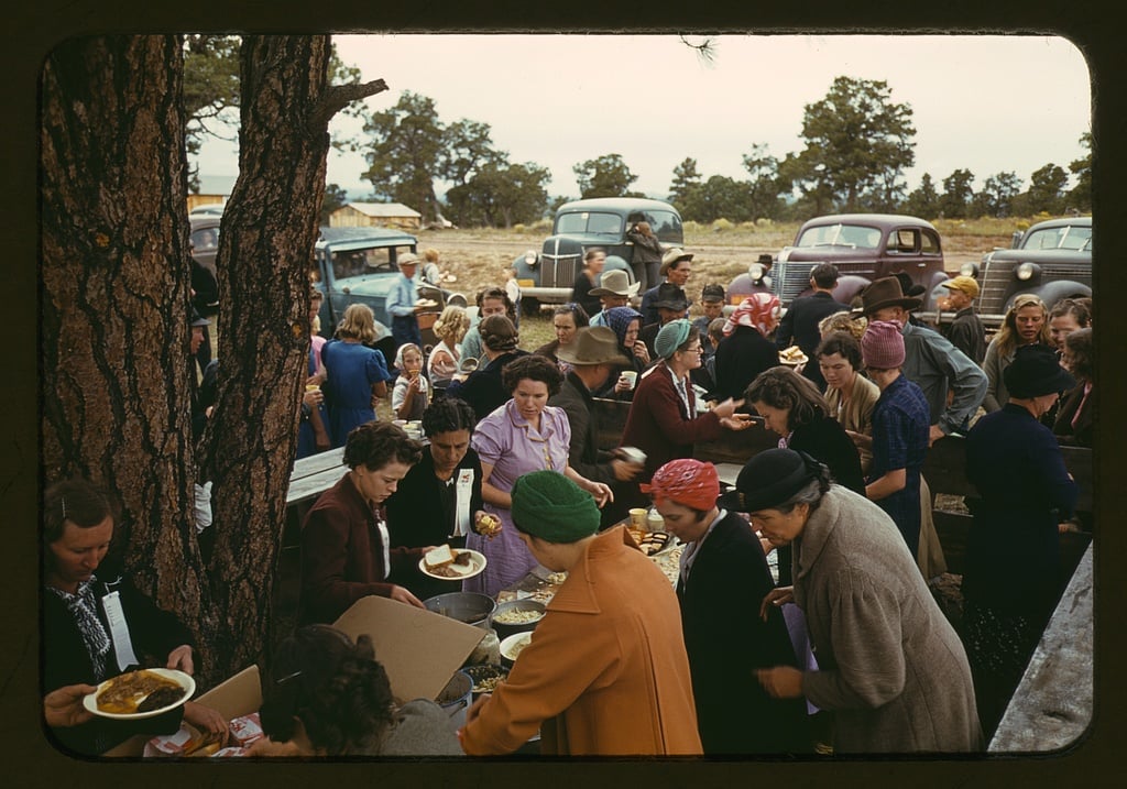 A large group of people, mostly women, gather outdoors around tables serving food. Some wear headscarves and coats, and vintage cars are parked in the background among trees. It appears to be a communal meal or event.