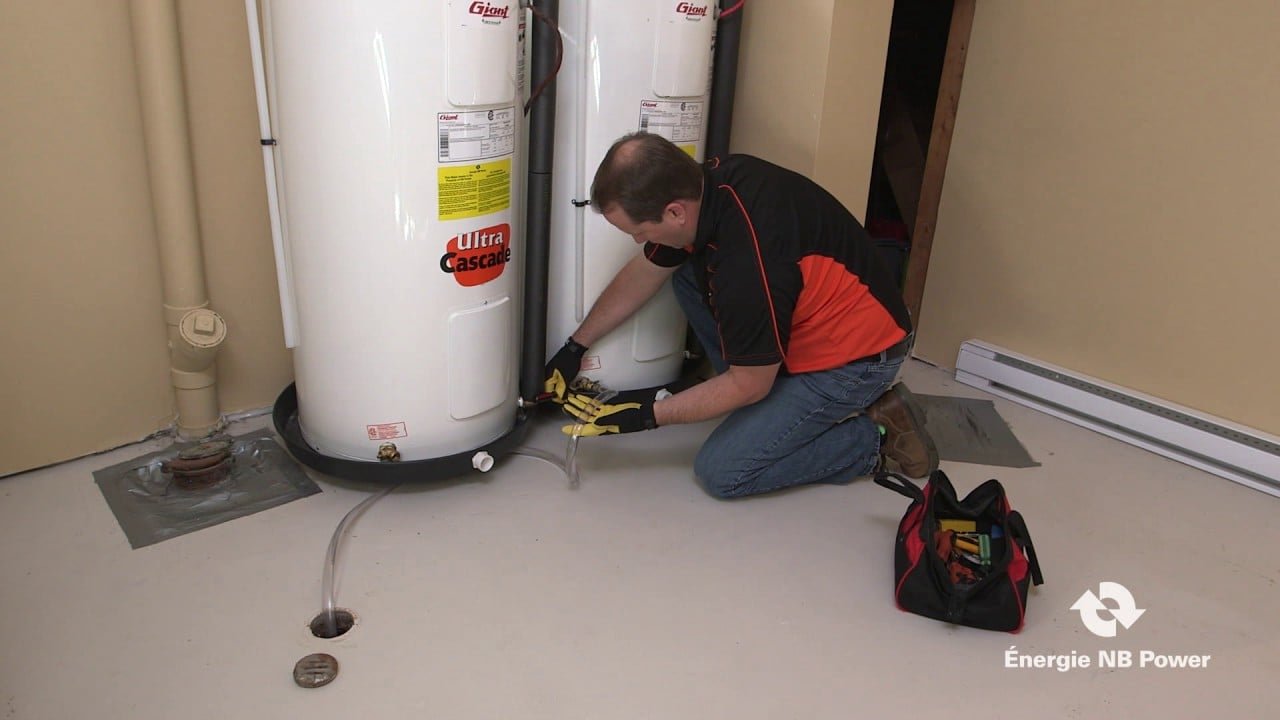 A person in gloves kneels on the floor, working on the bottom of a water heater with tools nearby. There are two water heaters and a tool bag is placed on the ground. The image includes an Énergie NB Power logo.