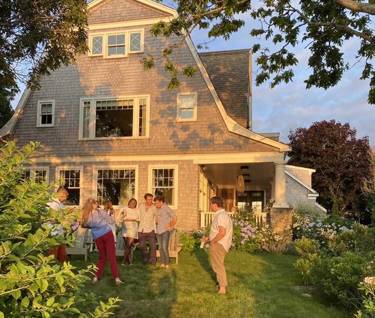 A group of people stand and talk in the garden of a large house with gray shingles, surrounded by greenery and flowers, in the warm light of late afternoon.