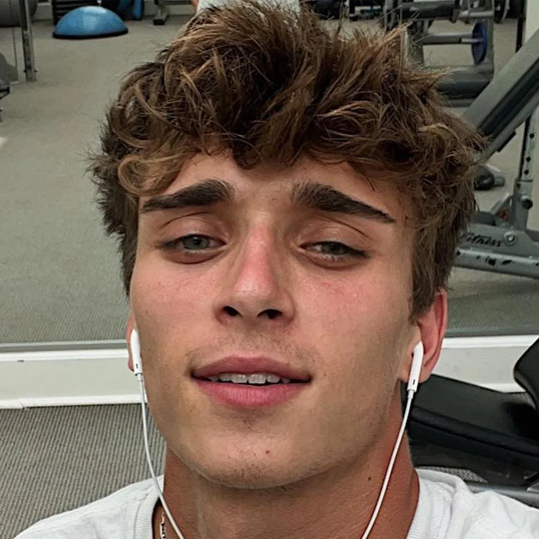 A young man with tousled brown hair and earbuds takes a selfie at the gym, with exercise equipment visible in the background. He has a slight smile and appears to be relaxed after a workout.