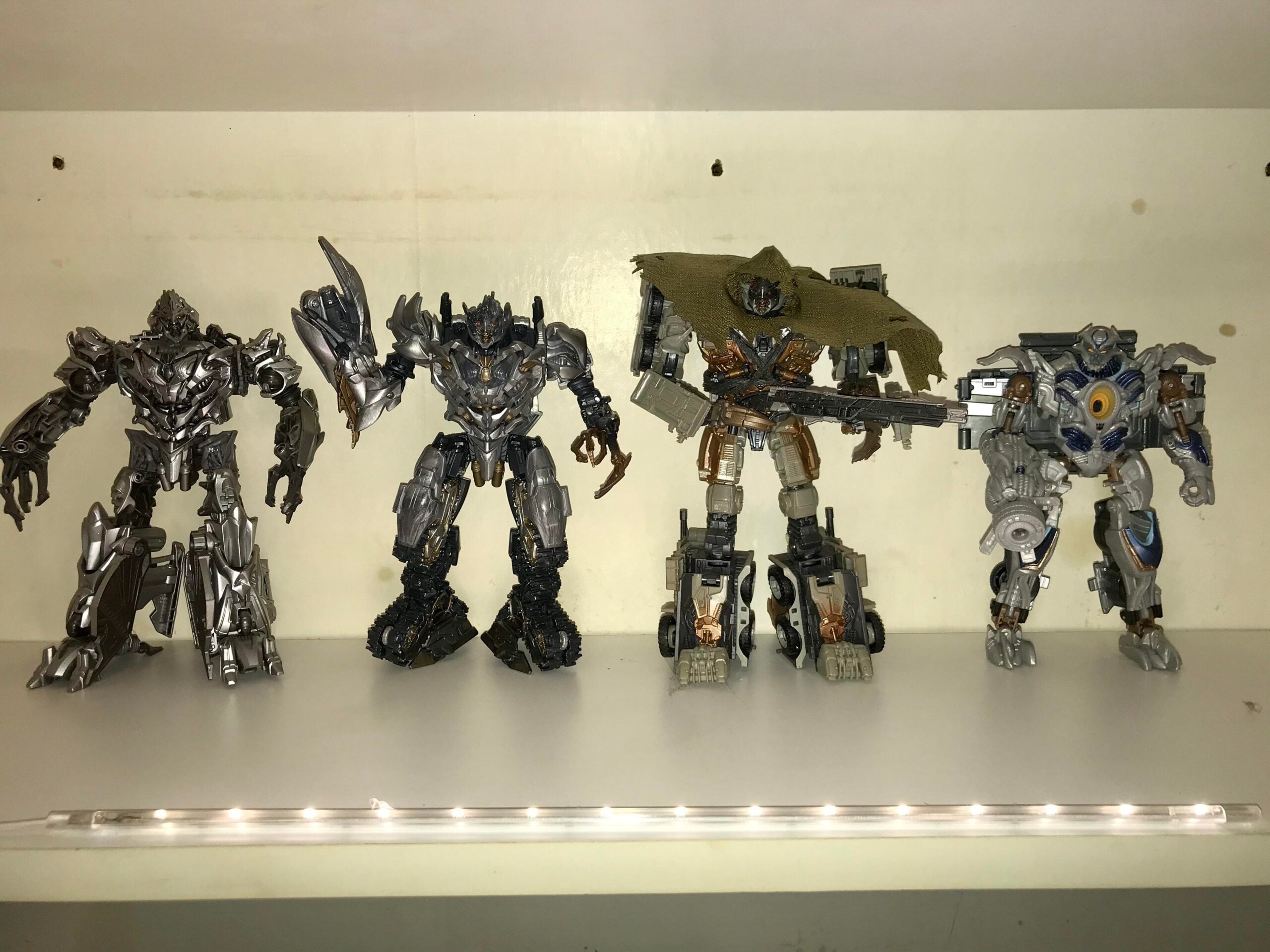 Four detailed Transformers action figures are displayed upright on a white shelf. The figures feature metallic armor and intricate designs, with one wearing a brown cape and another holding a weapon. The scene is brightly lit.