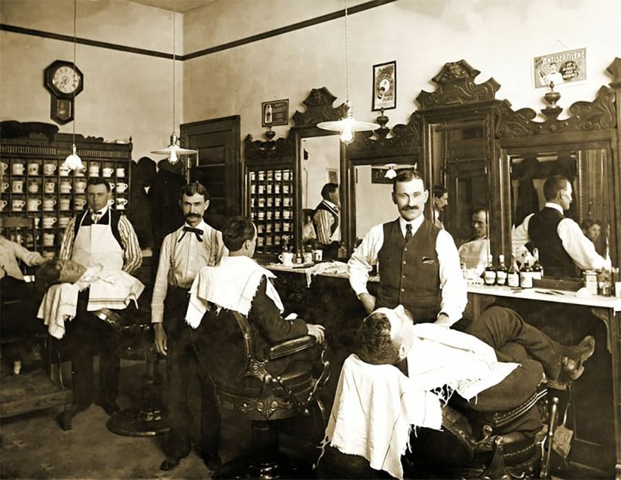 A vintage barbershop with barbers in vests and aprons attending to customers in barber chairs. The shop has ornate mirrors, shelves with bottles, and classic décor, creating a bustling, early 20th-century atmosphere.