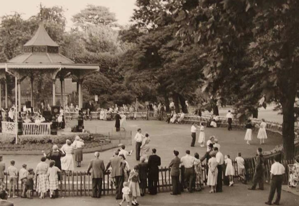 Black-and-white photo of people gathered in a park near a bandstand, watching dancers perform. Trees and benches line the background, and a crowd stands behind a low fence in the foreground.