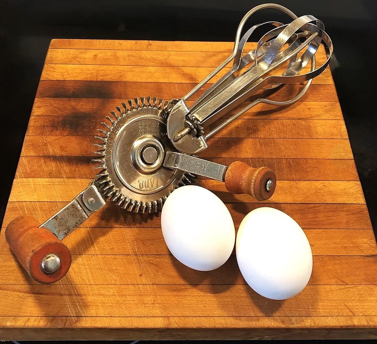 A hand-crank egg beater with wooden handles and two white eggs are placed on a wooden cutting board.
