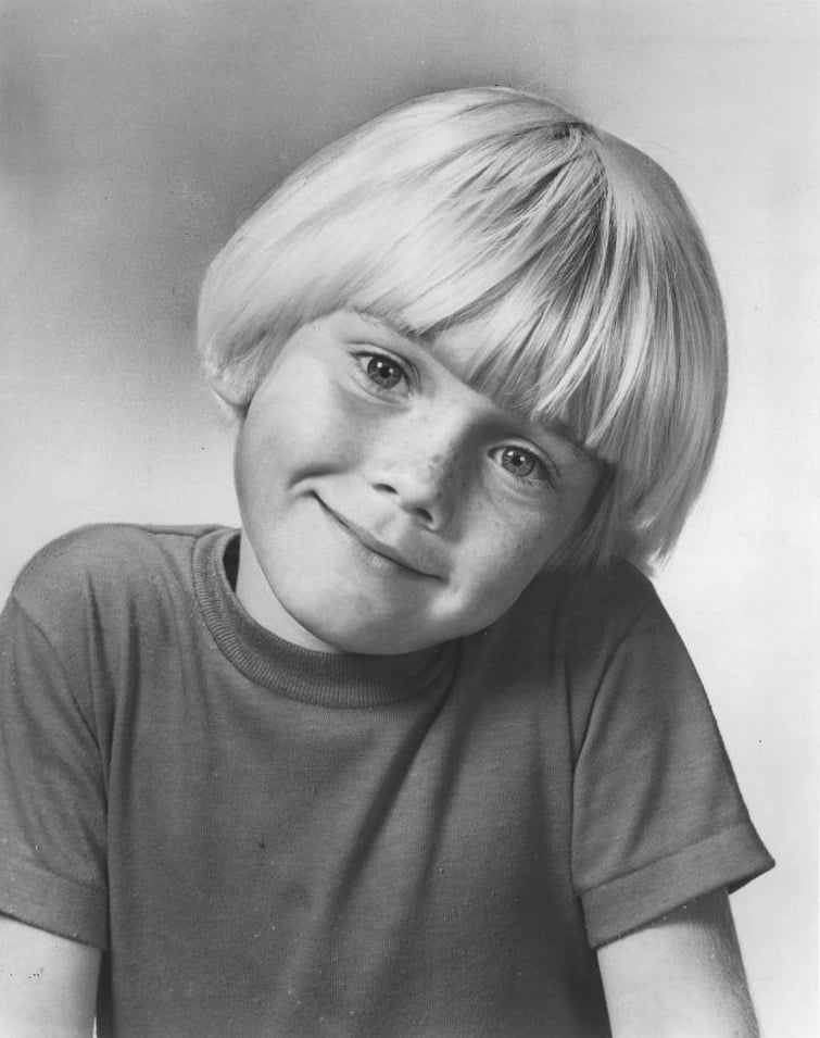 A young child with light blond hair in a bowl cut, wearing a plain short-sleeve shirt, smiles softly at the camera. The photo is black and white with a neutral background.