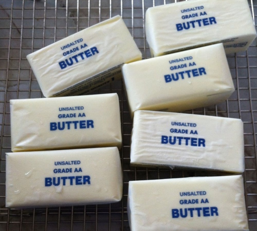 Seven sticks of unsalted Grade AA butter are arranged on a wire cooling rack. The butter sticks are still wrapped and have blue labeling on their wrappers.