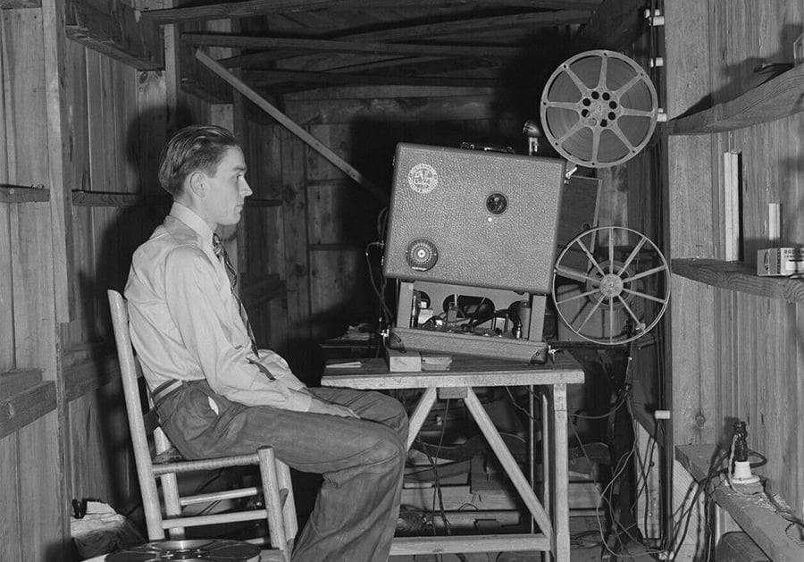 A man in a button-down shirt and tie sits on a wooden chair inside a wooden structure, operating a vintage film projector with large reels on a table in front of him.