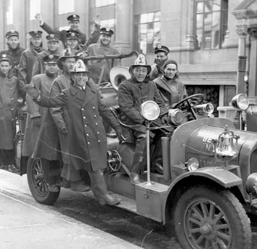 A group of firefighters in uniform pose and wave while riding an old-fashioned fire truck on a city street in front of a large building. The image appears to be from the early-to-mid 20th century.