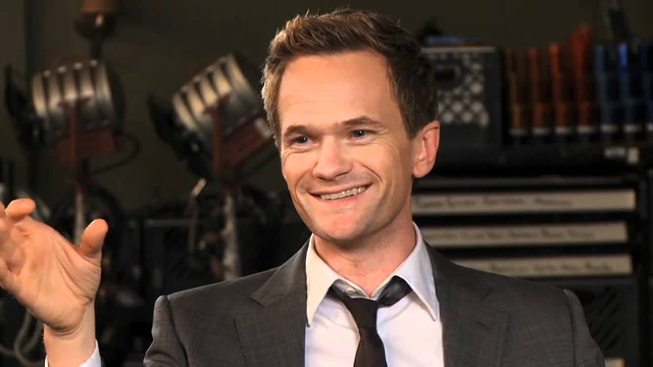 A smiling man in a gray suit and loosened tie gestures with his left hand while sitting indoors, with professional lighting equipment and shelves in the blurred background.