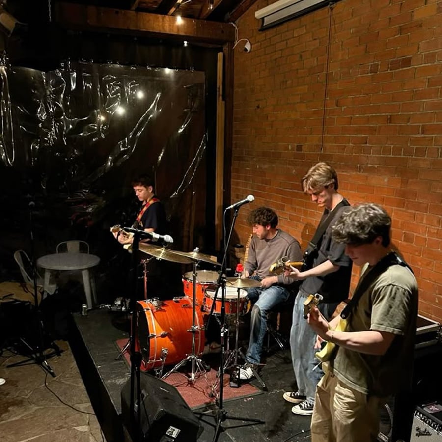 Four young men perform on a small indoor stage; one plays drums while the others play electric guitars. The setting features exposed brick walls and dim lighting, creating an intimate atmosphere.