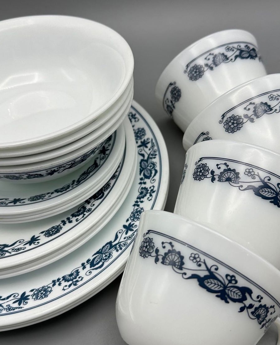A close-up of stacked white dishes and bowls with a matching set of white cups, all featuring a blue floral and vine pattern around the rim, set against a gray background.